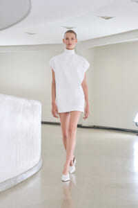 Runway model wearing a white minimalist high-neck tunic dress with cap sleeves, walking in a bright studio.