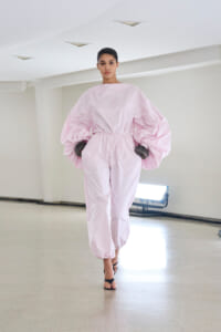 Model walks a runway wearing a pale pink, oversized jumpsuit with dramatic balloon sleeves and an elastic waist, paired with black sandals in a minimalist studio.