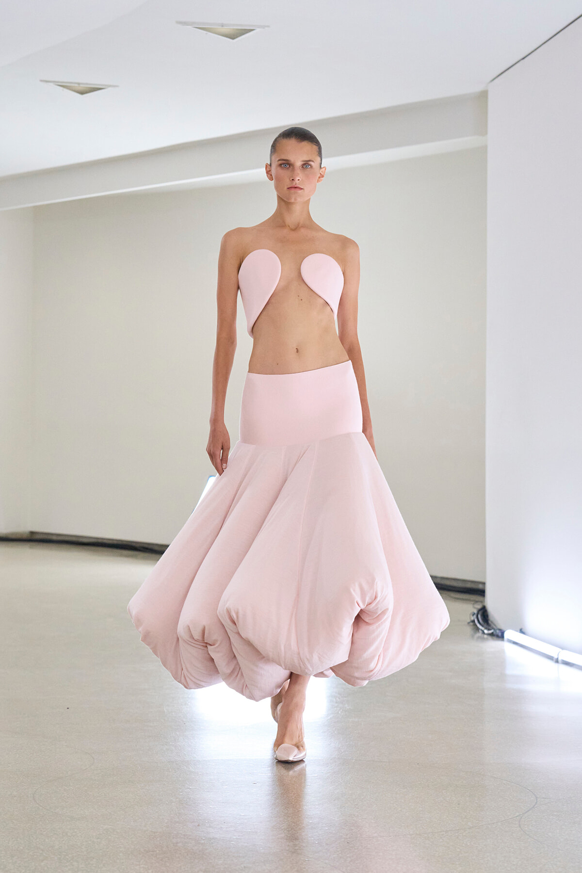 Female model walking in a pale pink strapless dress with a voluminous bubble skirt in a white studio.