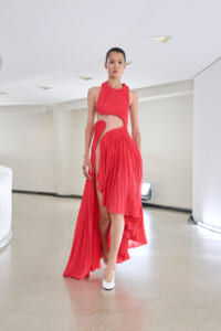 Model in a red pleated halter dress with cutouts walking down a minimalist white hallway runway with white heels.