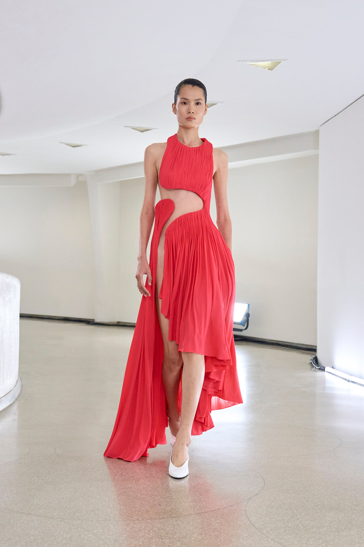 Model in a red pleated halter dress with cutouts walking down a minimalist white hallway runway with white heels.