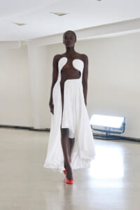 Model walking a runway in a white pleated gown with sculptural chest and waist cutouts, paired with red heels.