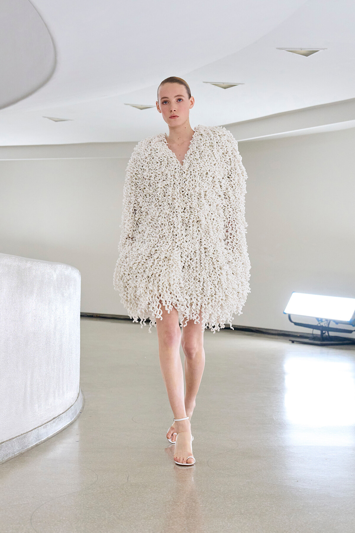 Model walking in a white, oversized textured dress made of looped fabric in a minimalist studio.