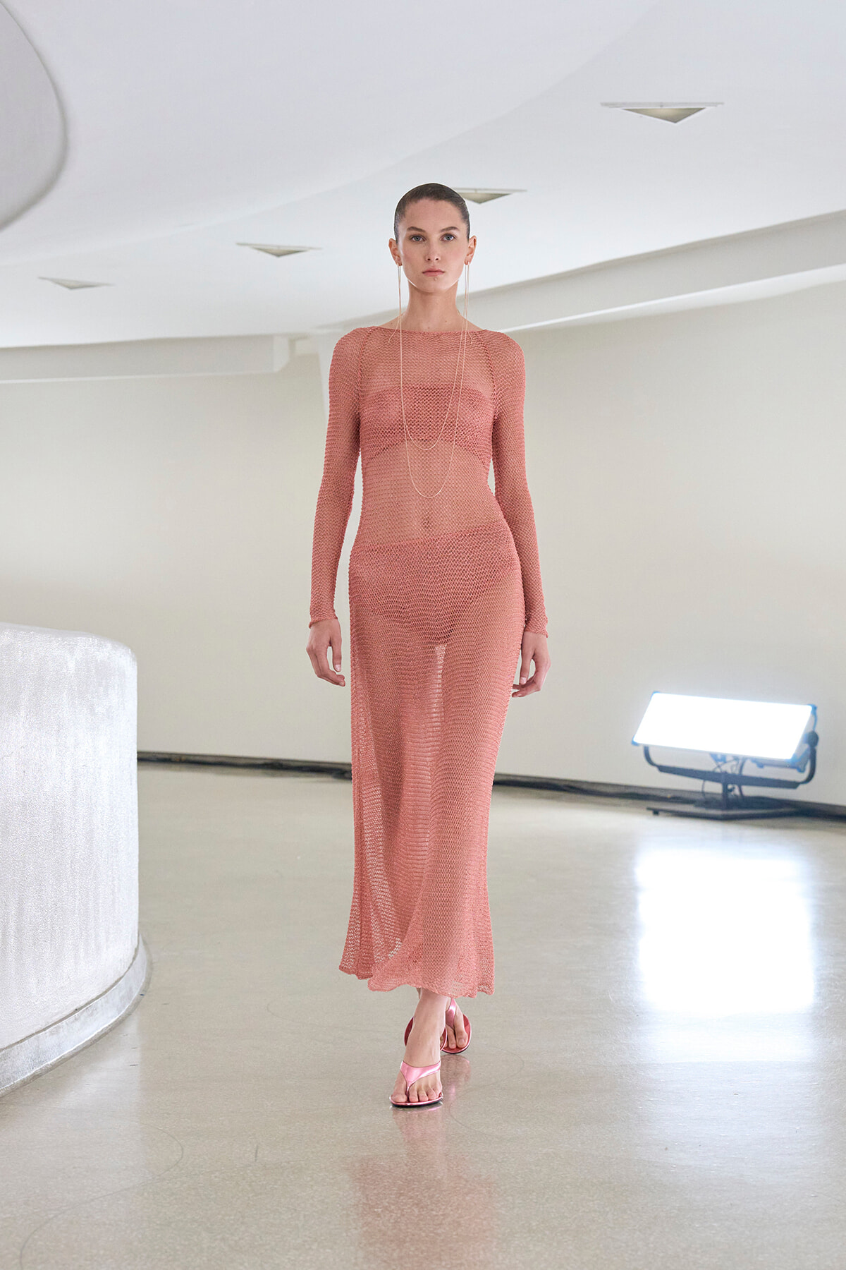 Model walking in a peach sheer knit maxi dress with long sleeves, paired with pink sandals in a bright, minimalist studio.