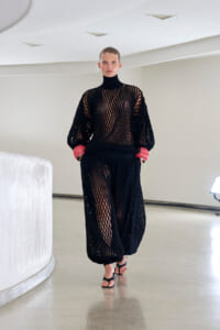 Model in a black mesh knit turtleneck top and wide-leg pants with red cuffs, walking in a bright studio.