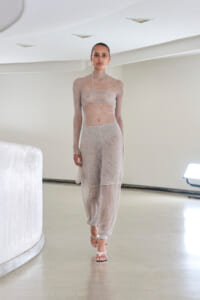 Model walking in a sheer, knit two-piece outfit (turtleneck top and wide-leg pants) with sandal heels in a bright, minimalist studio.