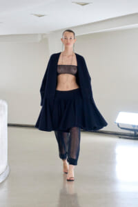 Model walking in a navy oversized coat over a black mesh bandeau, with a voluminous navy skirt and mesh pants, wearing open-toe heels in a minimalist studio setting.