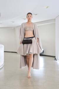 Runway model in a beige, flowing coat over a mesh bandeau and sculpted mini skirt, with a small black waist bag and nude heels.