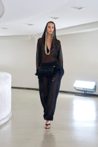 Model walks a runway in a black sheer hooded jumpsuit with a black waist bag and long beige necklace accessories.