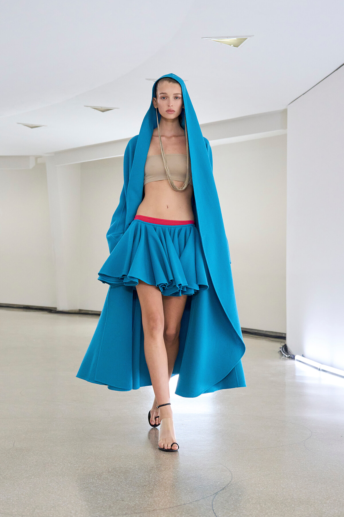 Model walking in a bright blue hooded cape over a beige bandeau top and a voluminous turquoise skirt with a red waistband, in a minimalist white studio.