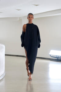 Model walks in a black, oversized one-shoulder dress with a high slit, wearing a long necklace and strappy heels in a minimalist studio setting.