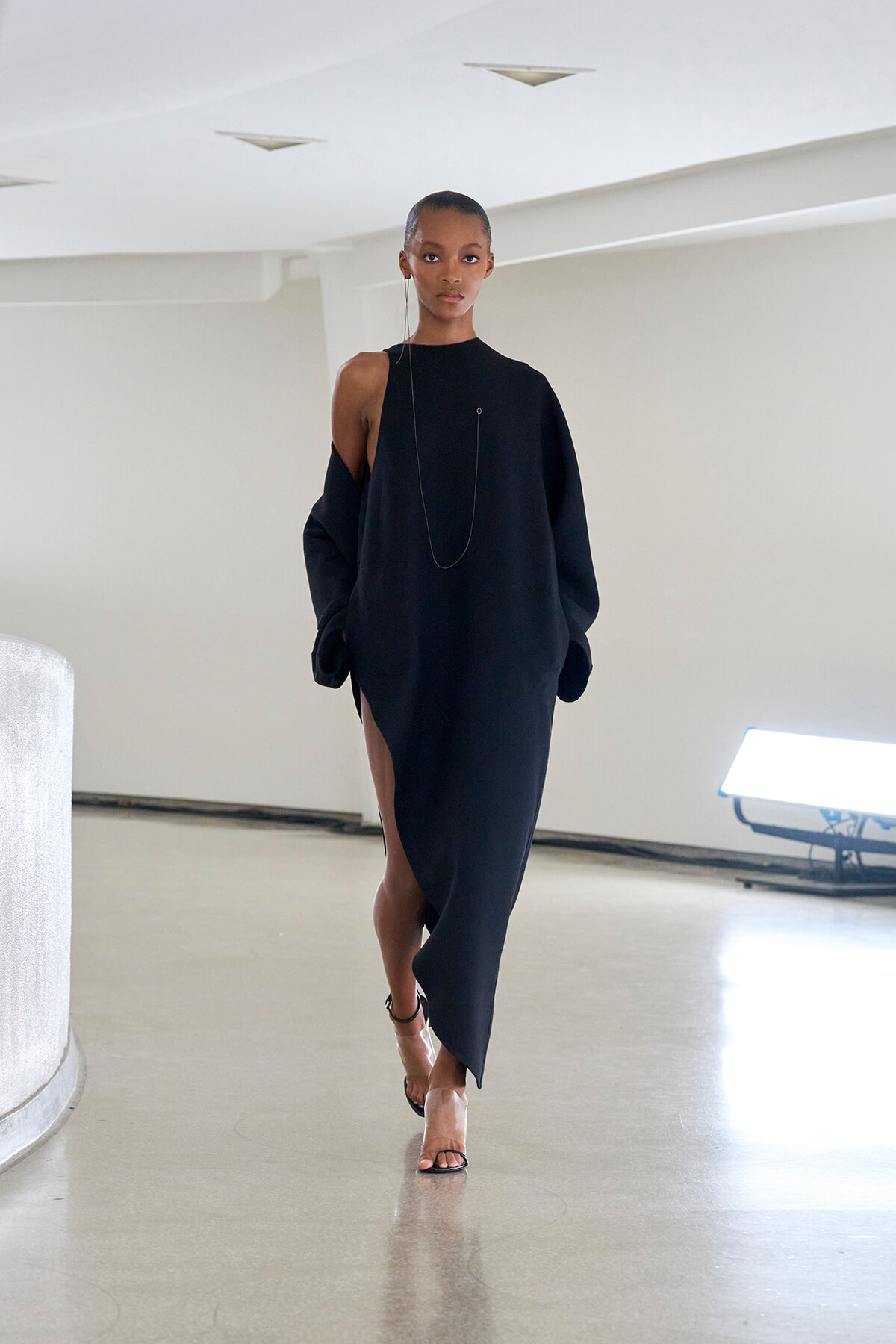Model walks in a black, oversized one-shoulder dress with a high slit, wearing a long necklace and strappy heels in a minimalist studio setting.