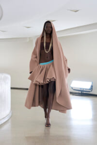Model walking a runway in a taupe hooded cape over a brown bandeau top and a layered ruffled skirt with a turquoise waistband.