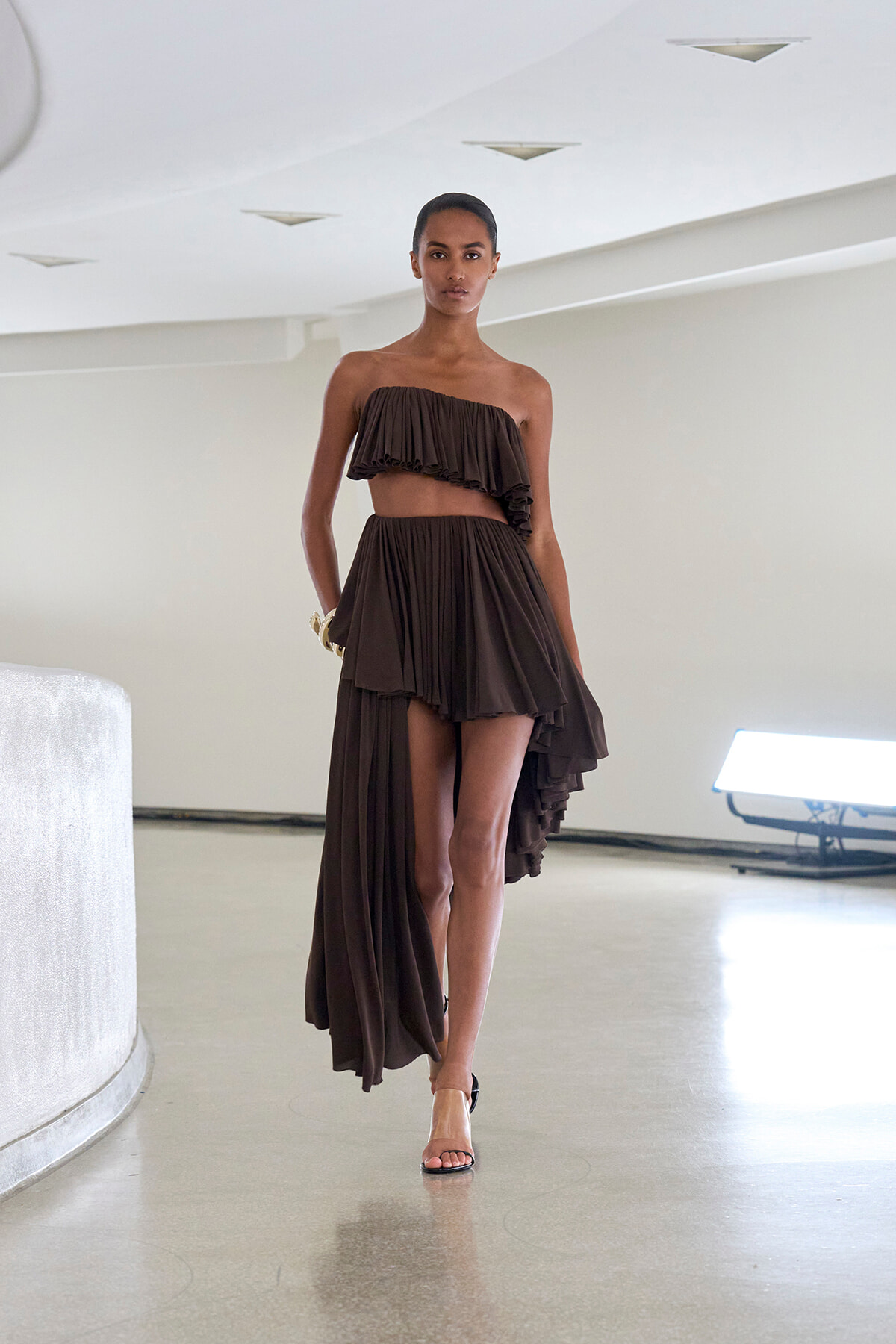 Model walks the runway in a dark brown one-shoulder, ruffled crop top paired with a matching draped, high-low skirt.