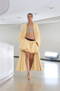 Model walking in a pale yellow cape-style coat over a beige bandeau and layered mini skirt on an indoor runway, wearing beige sandals.