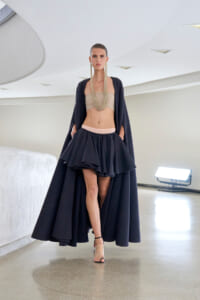 Model walking a runway in a navy cape and voluminous layered skirt over a beige bandeau, accented with a long multi-strand necklace.