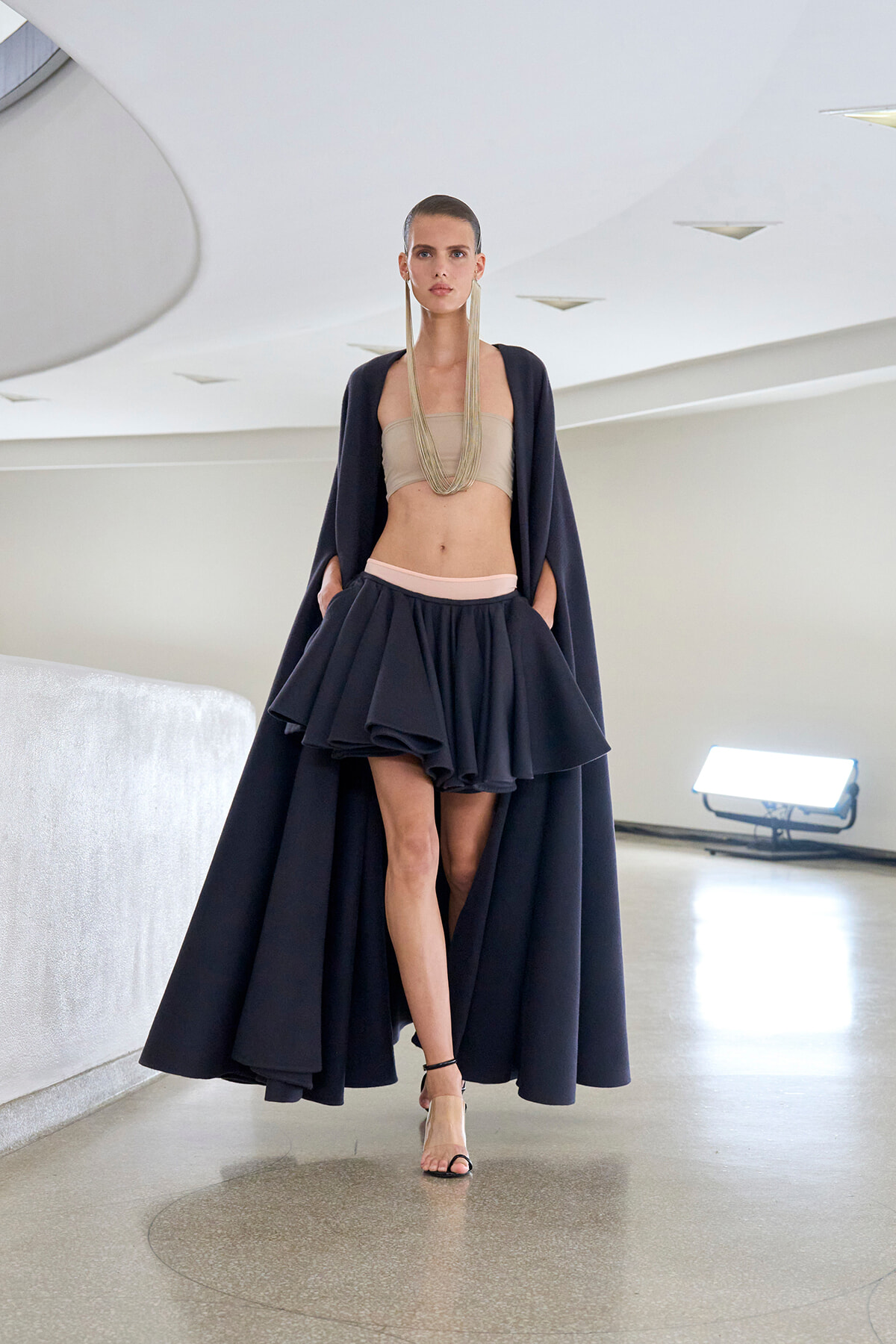 Model walking a runway in a navy cape and voluminous layered skirt over a beige bandeau, accented with a long multi-strand necklace.
