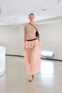 Model walking a runway in a beige bandeau top and pale pink layered skirt with a brown waistband, carrying a small black crossbody bag in a bright studio setting.