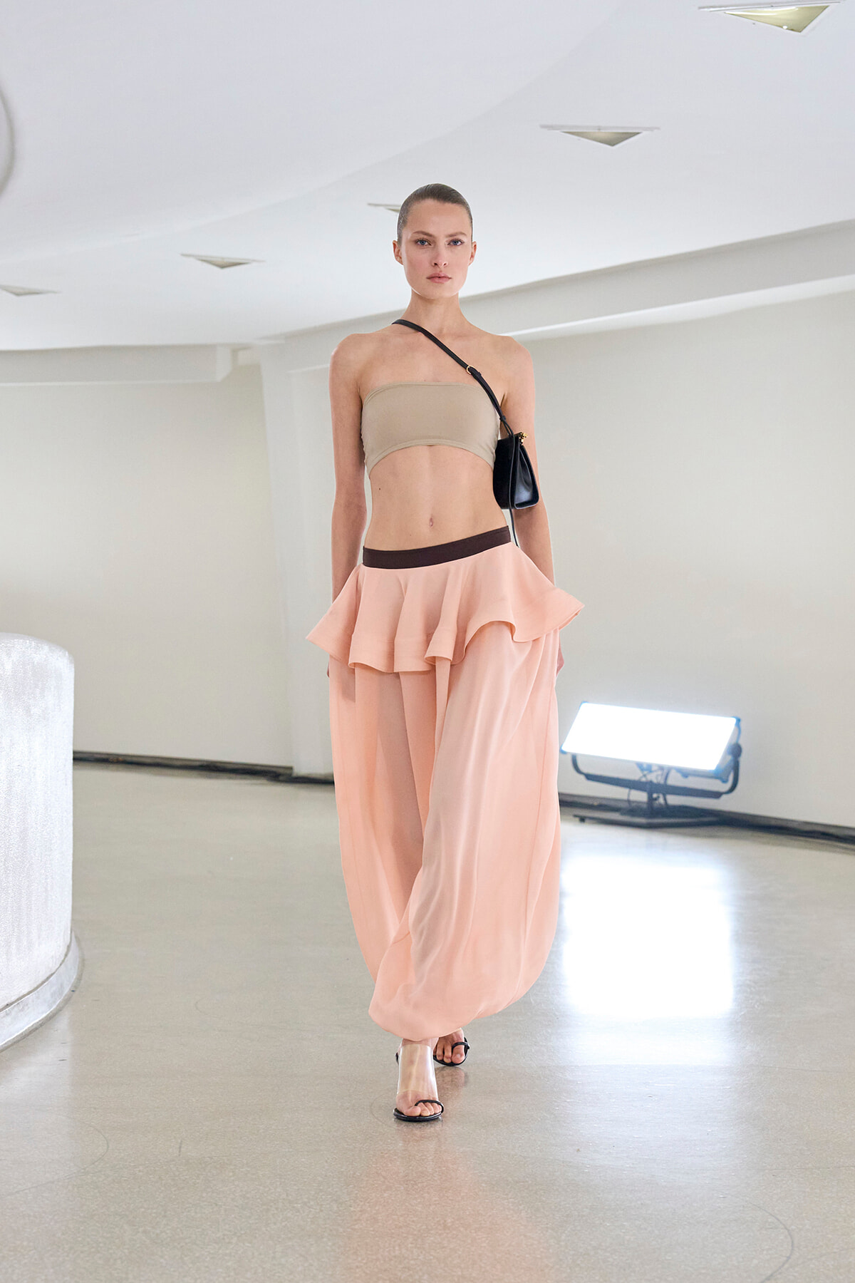 Model walking a runway in a beige bandeau top and pale pink layered skirt with a brown waistband, carrying a small black crossbody bag in a bright studio setting.