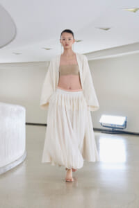 Runway model in an ivory flowing skirt and open cardigan, beige bandeau top, walking a minimalist studio runway.
