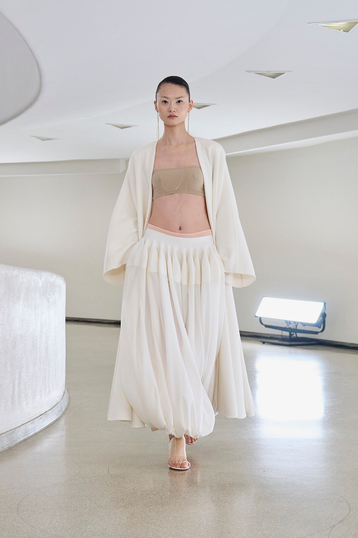 Runway model in an ivory flowing skirt and open cardigan, beige bandeau top, walking a minimalist studio runway.