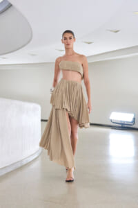 Model wearing a one-shoulder beige two-piece dress with a layered high-low skirt walking down a modern, white-curved hallway.