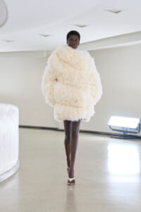 Model walks a minimalist runway in an oversized cream fur coat, neutral space and soft lighting.