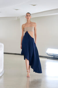 Model walking a runway in a beige bandeau top and navy asymmetric wrap skirt with long dangling earrings on a minimalist setting.