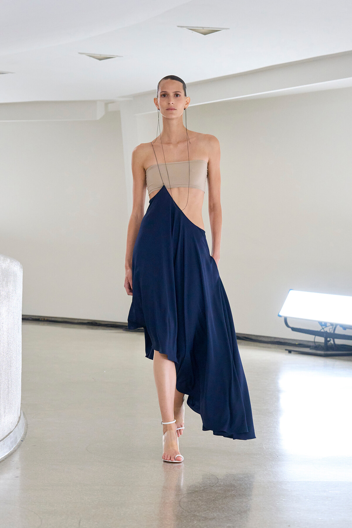 Model walking a runway in a beige bandeau top and navy asymmetric wrap skirt with long dangling earrings on a minimalist setting.