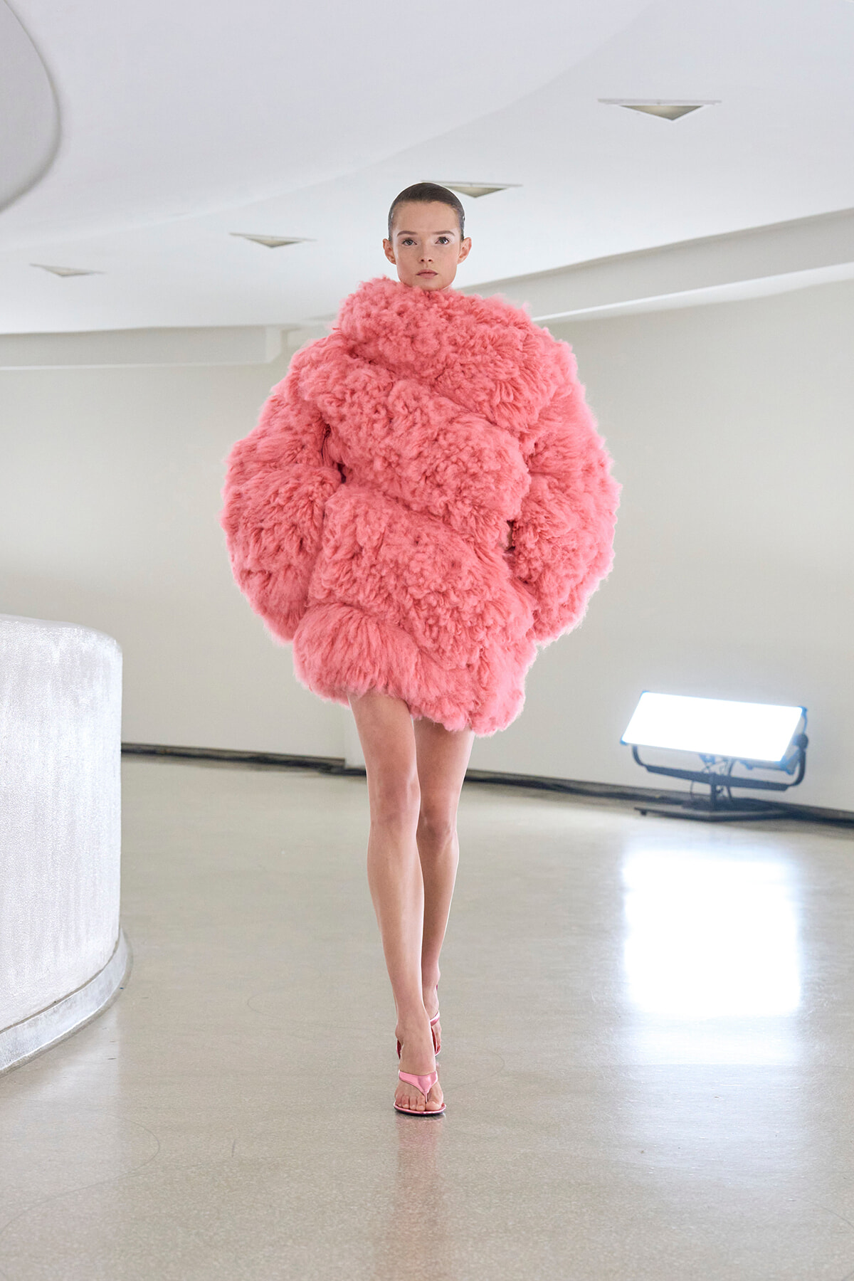 Model walking a runway in an oversized pink fluffy faux-fur coat with pink sandals in a bright, minimalist space.
