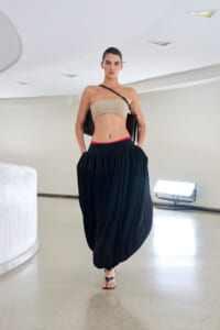 Model walking in a neutral studio wearing a beige bandeau top and a voluminous black draped skirt with sandals and a shoulder bag.