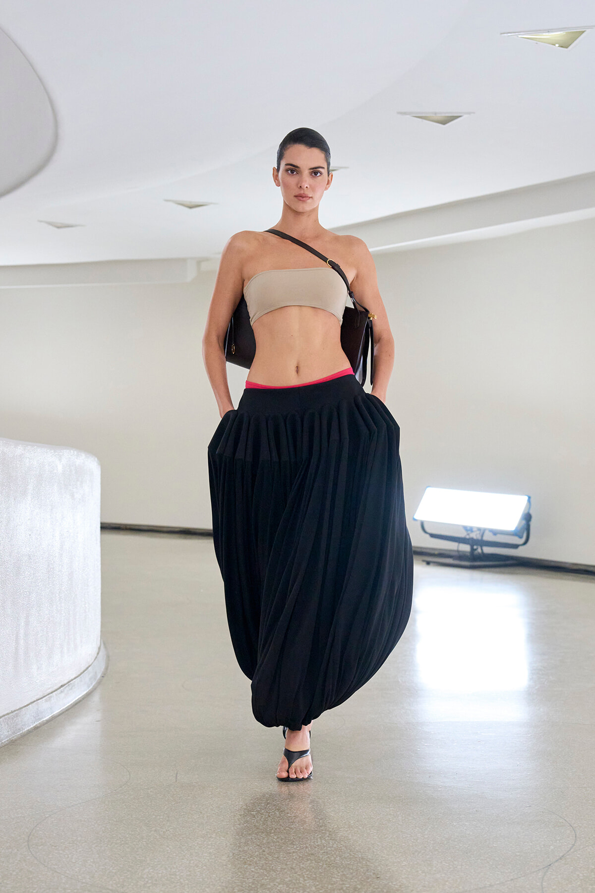 Model walking in a neutral studio wearing a beige bandeau top and a voluminous black draped skirt with sandals and a shoulder bag.