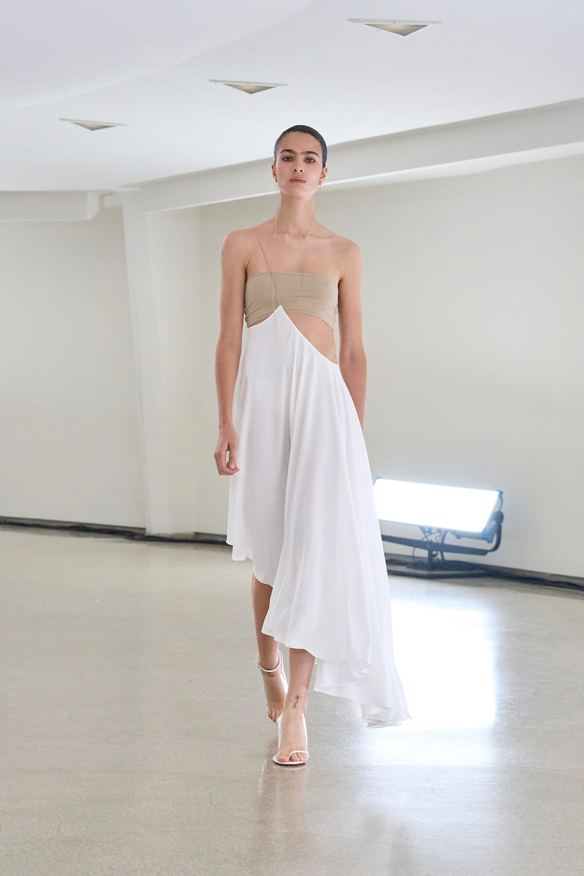 Model walks the runway in a beige bandeau top and white draped, asymmetrical dress in a bright studio setting.
