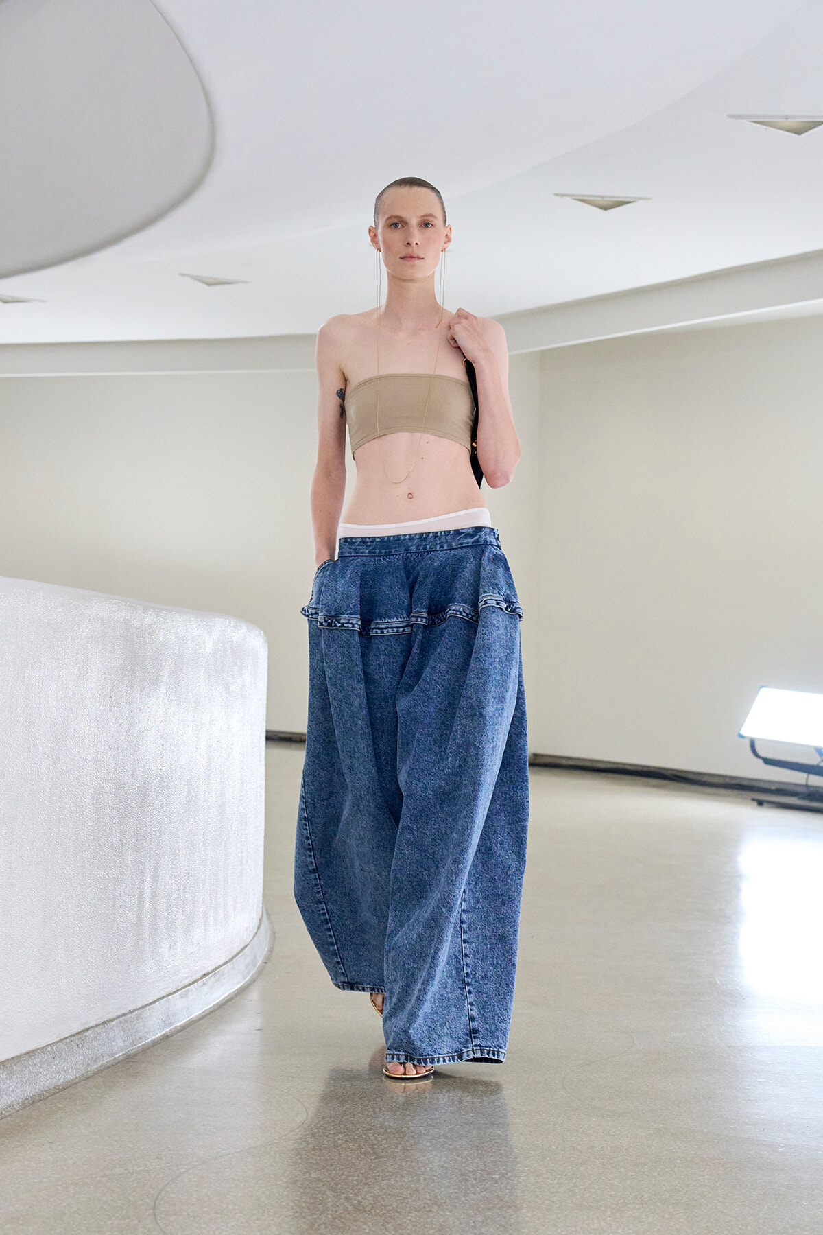 Model wearing a tan bandeau top and ultra-wide denim pants, walking in a bright white studio space.