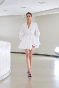 Model walking in a white V-neck dress with a layered gold necklace in a minimalist studio setting.