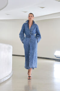 Model wearing a long denim jumpsuit with hands in pockets, walking barefooted in a bright, minimalist space.