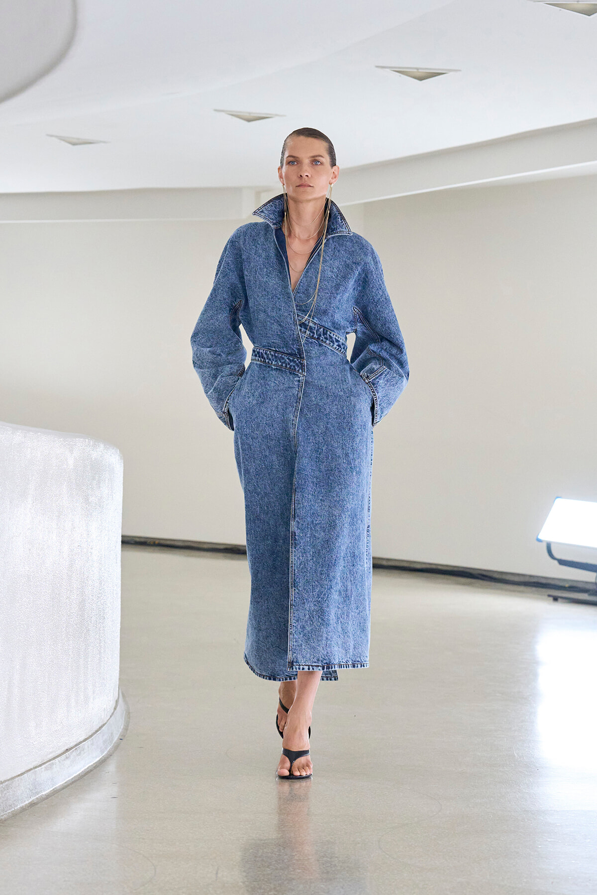 Model wearing a long denim jumpsuit with hands in pockets, walking barefooted in a bright, minimalist space.