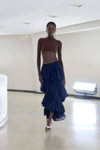 Model wearing a sheer brown long-sleeve crop top with a layered navy ruffle skirt in a bright studio setting, standing confidently on a polished floor with white walls and soft lighting.