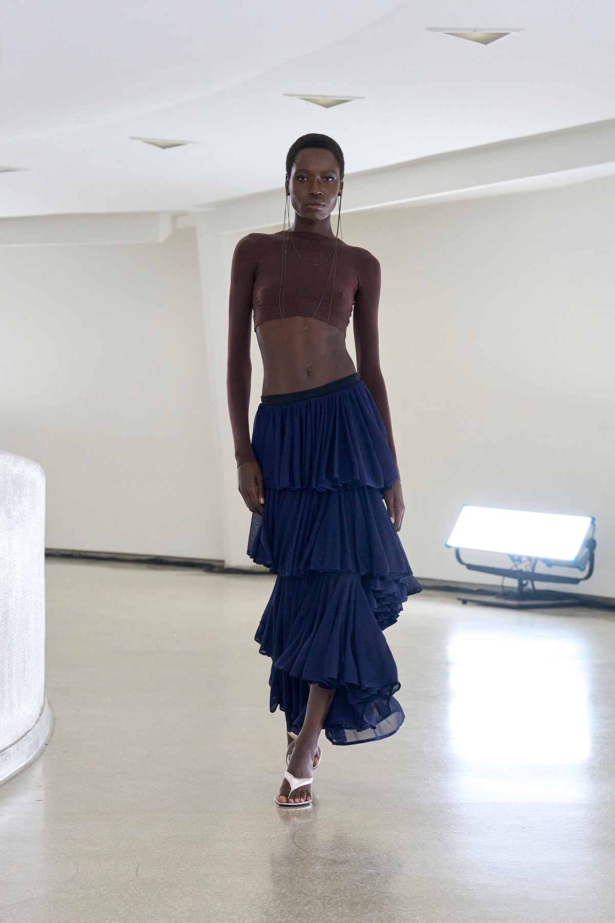 Model wearing a sheer brown long-sleeve crop top with a layered navy ruffle skirt in a bright studio setting, standing confidently on a polished floor with white walls and soft lighting.
