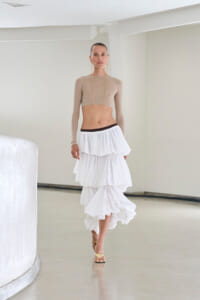 Model wearing a beige long-sleeve cropped top and a white multi-tiered skirt walking in a bright studio setting, minimalist background.