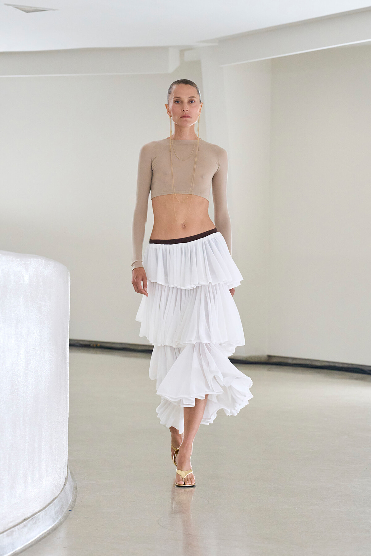 Model wearing a beige long-sleeve cropped top and a white multi-tiered skirt walking in a bright studio setting, minimalist background.