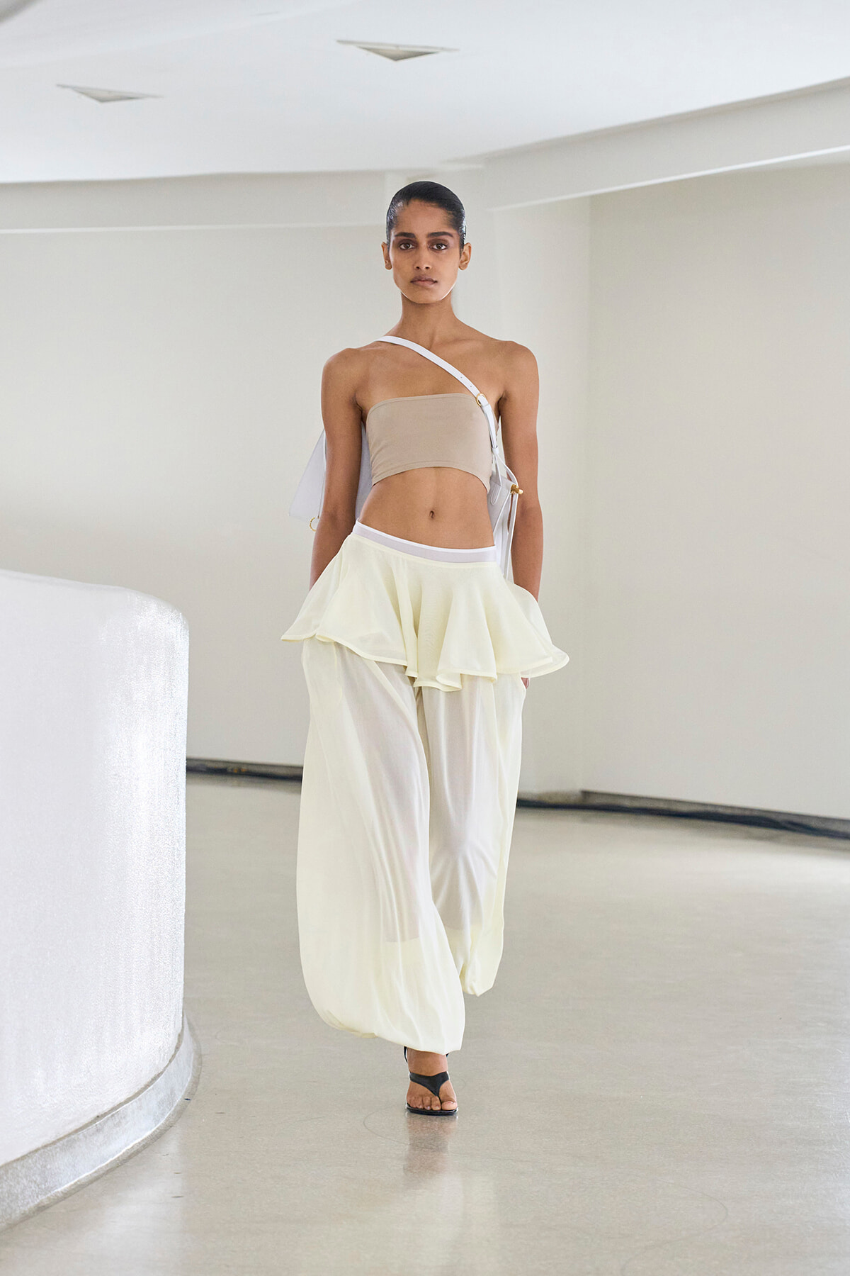 Model walking a runway in a beige bandeau top and flowing white layered trousers in a minimalist setting.