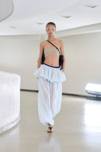 Model walking a runway in a beige bandeau top, light blue layered pants and black crossbody bag in a bright, minimal setting.