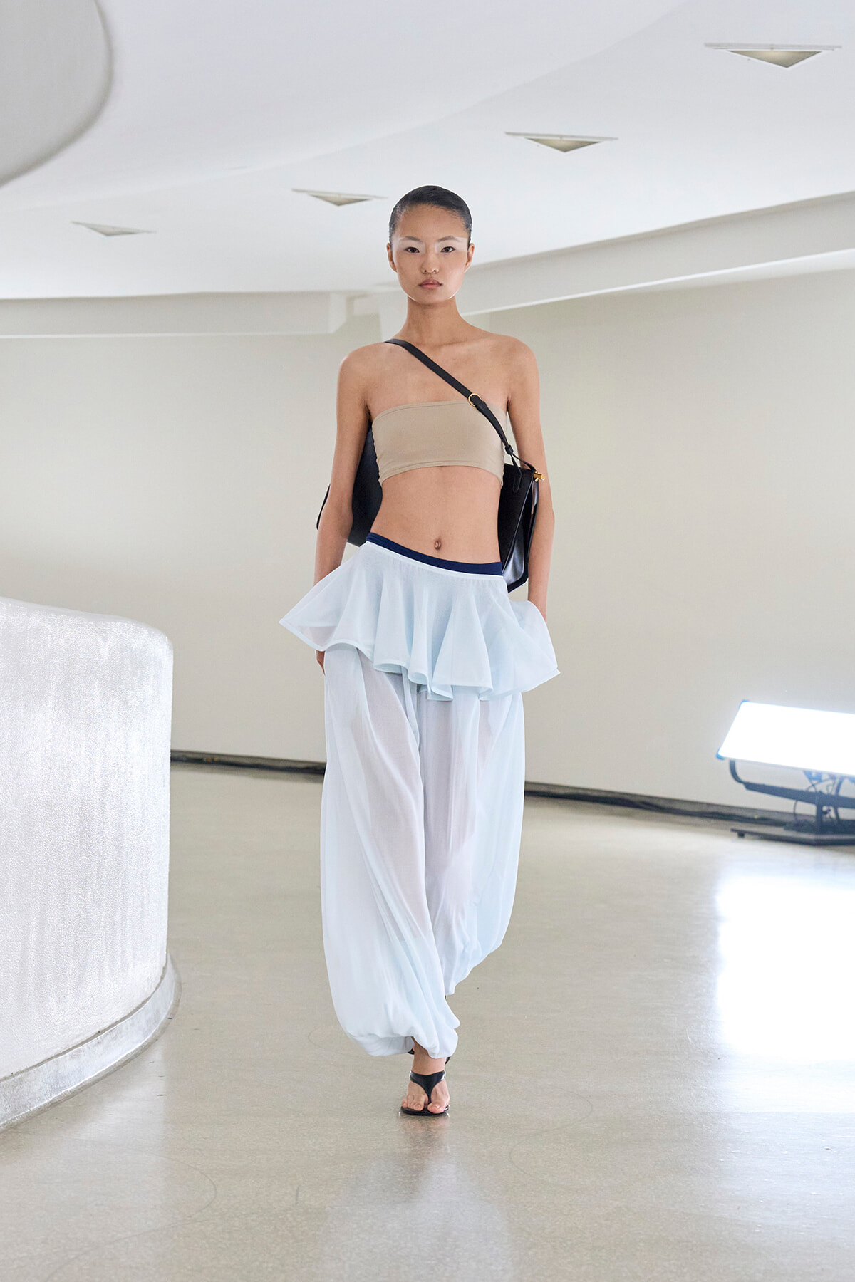Model walking a runway in a beige bandeau top, light blue layered pants and black crossbody bag in a bright, minimal setting.