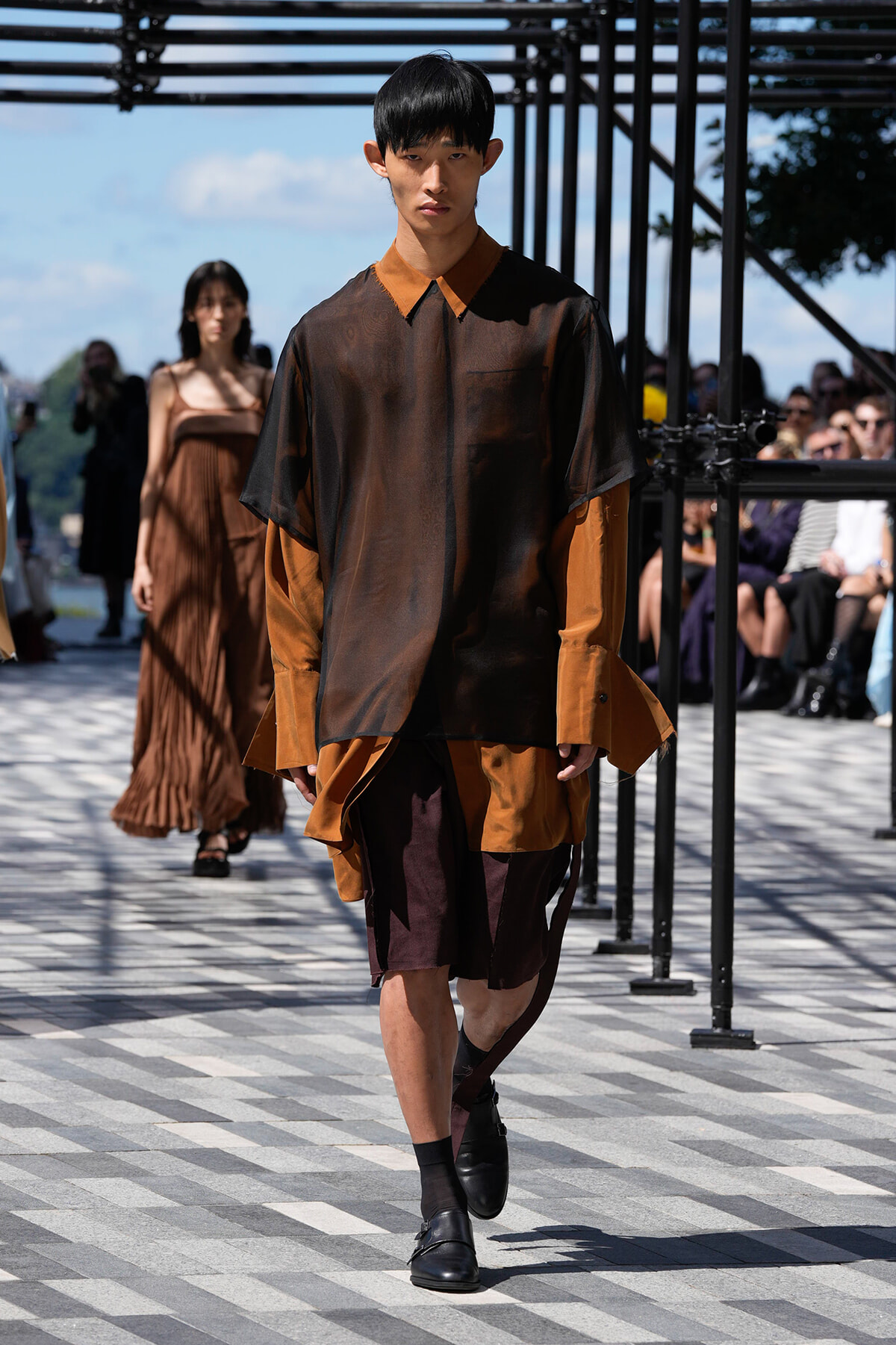 Male model walks a runway in a sheer brown-black top with orange collar, layered over rust-colored garments, outdoors with spectators in background.