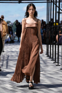 Model walking on an outdoor runway in a brown pleated dress with a slit, sandals, and a neutral expression.