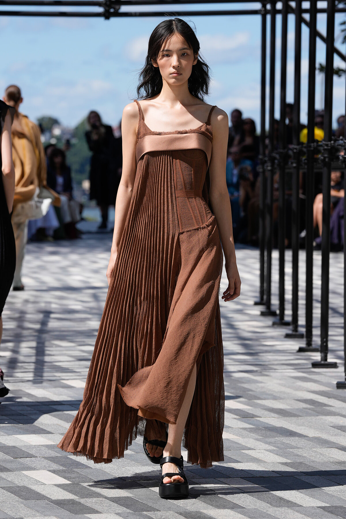 Model walking on an outdoor runway in a brown pleated dress with a slit, sandals, and a neutral expression.