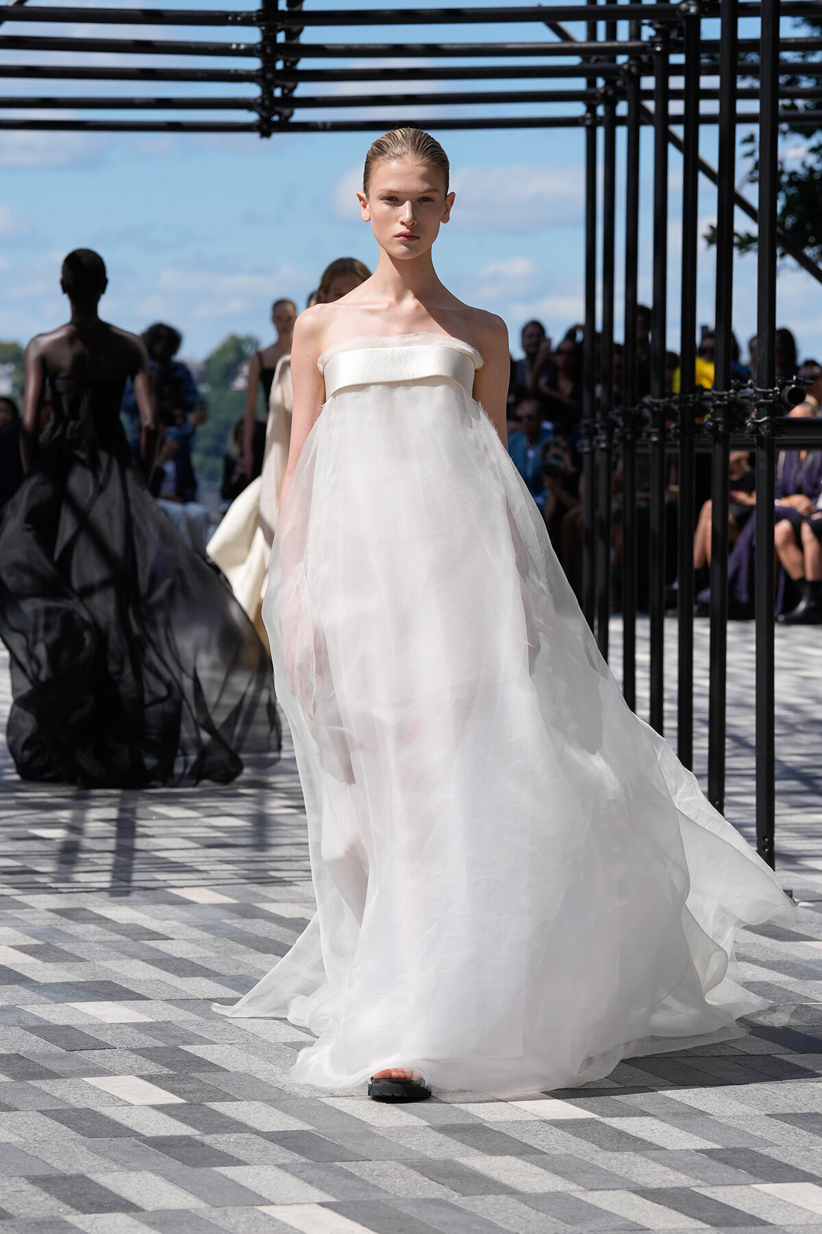 Model walks a fashion runway in a white strapless, floor-length gown with a sheer skirt, under a black metal framework with a blue sky backdrop and audience in the background.