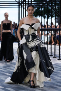 Model walks a runway in a dramatic black-and-white off-shoulder gown with abstract paint-like patterns, spectators behind.