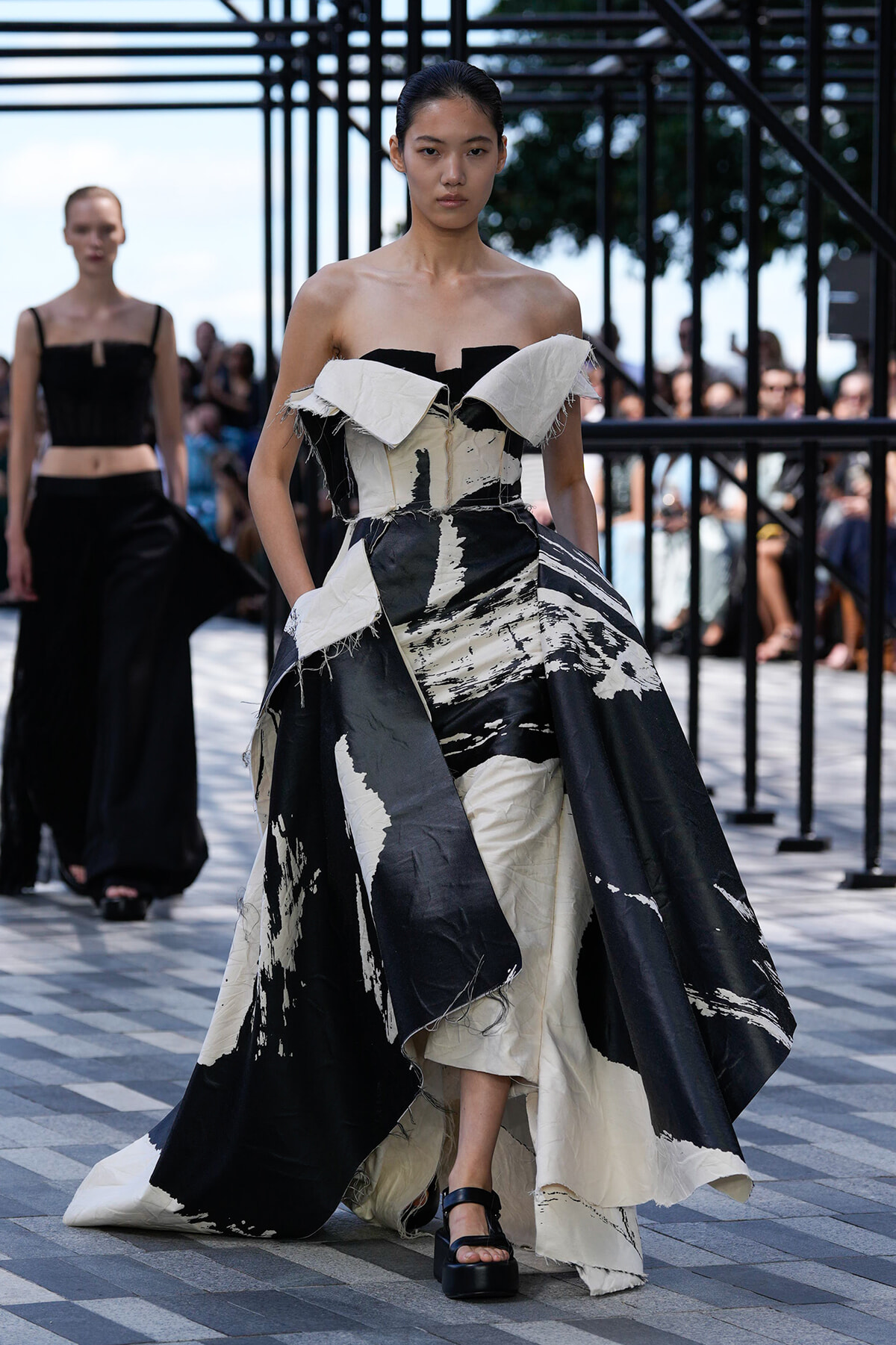 Model walks a runway in a dramatic black-and-white off-shoulder gown with abstract paint-like patterns, spectators behind.